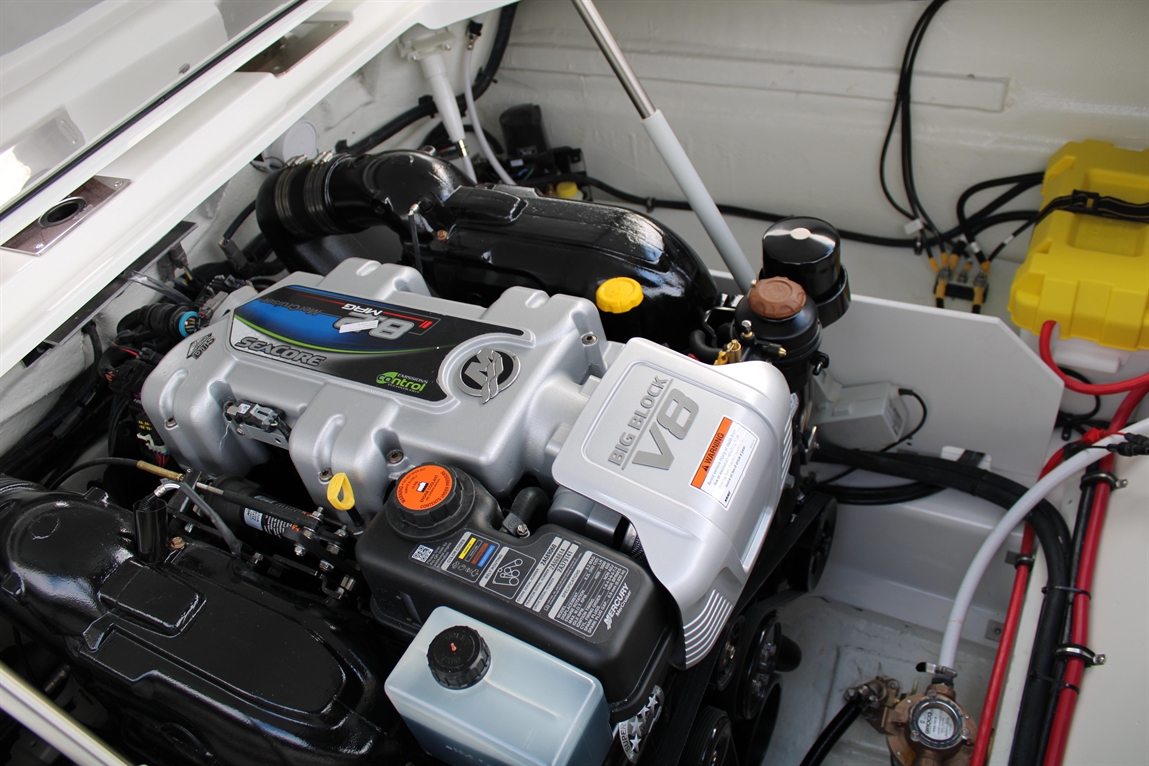 A big-block V8 engine viewed from above, featuring various hoses, caps, and a yellow reservoir in a boat engine compartment.